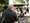 Ein Mann mit grauen Haaren und Rucksack steht im Vordergrund und blickt auf eine Warteschlange von Menschen unter einem schwarzen Sonnenschirm. Im Hintergrund sitzen Gäste an Tischen eines Außenbereichs.