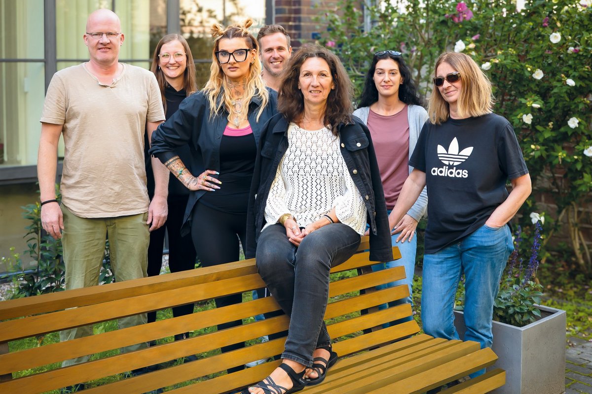 Sieben Personen posieren lächelnd für ein Gruppenfoto im Freien. Eine Frau sitzt auf einer Holzbank im Vordergrund, während die anderen sechs hinter ihr stehen. Im Hintergrund sind Pflanzen und ein Gebäude zu erkennen.