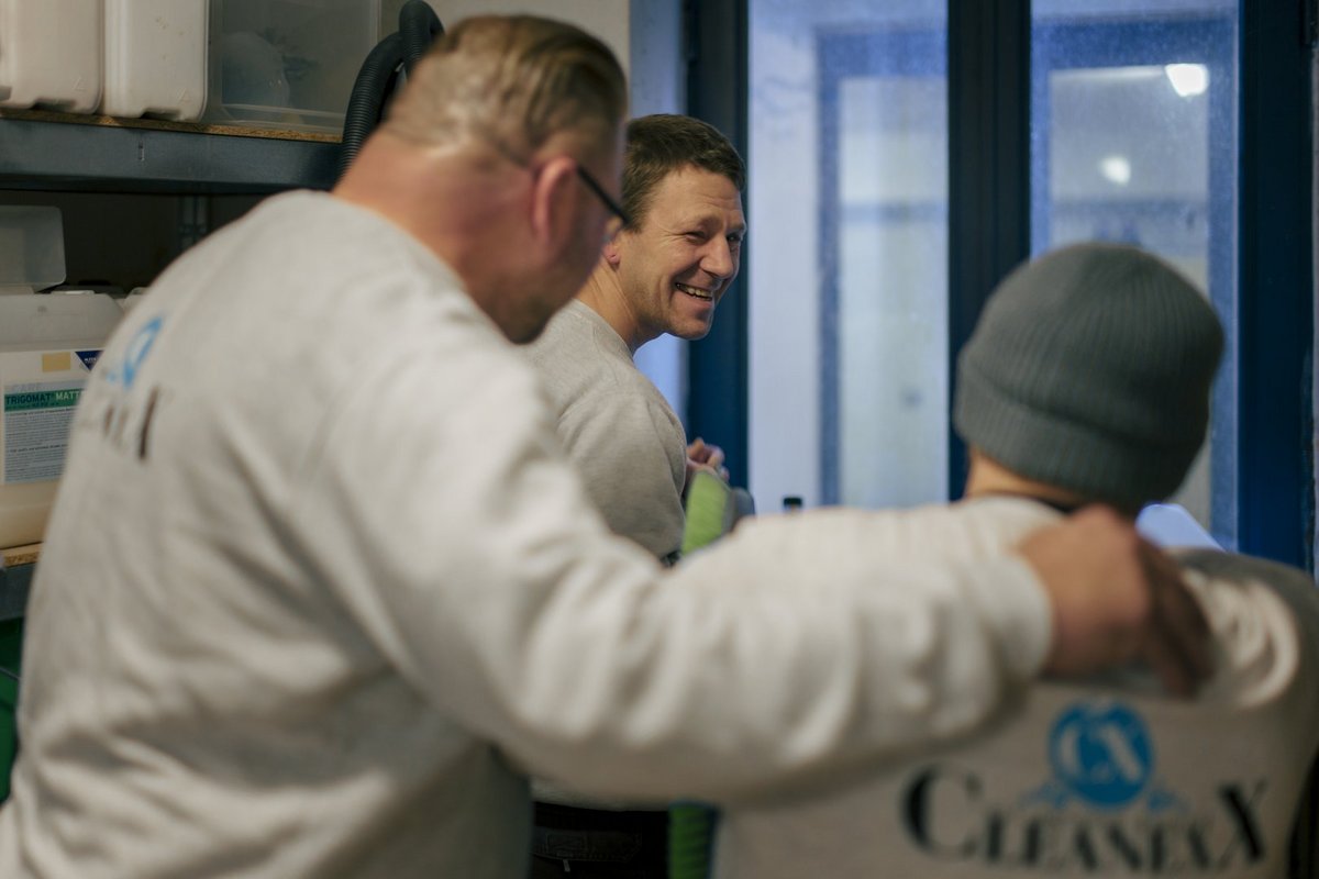 Drei Männer in weißen „CLEANEX"-Sweatshirts stehen in einer Gruppe. Ein Mann im Hintergrund lächelt herzlich, während ein anderer im Vordergrund dem dritten Mann mit der grauen Mütze freundschaftlich auf die Schulter legt. Die Szene zeigt einen Moment der Kollegialität und Freude.