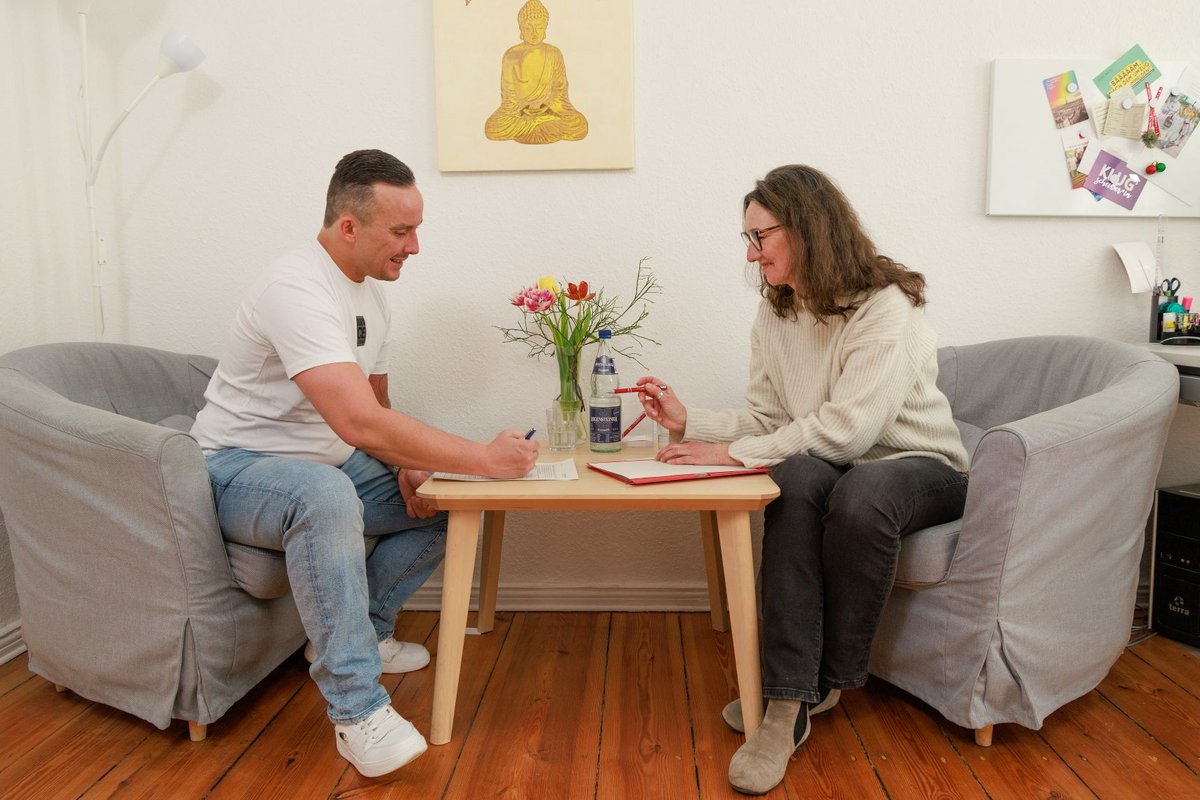Ein Mann und eine Frau sitzen sich in grauen Sesseln gegenüber und führen ein Gespräch. Zwischen ihnen steht ein kleiner Holztisch mit Blumen und Wasser. Die Szene wirkt wie eine Beratungssitzung.