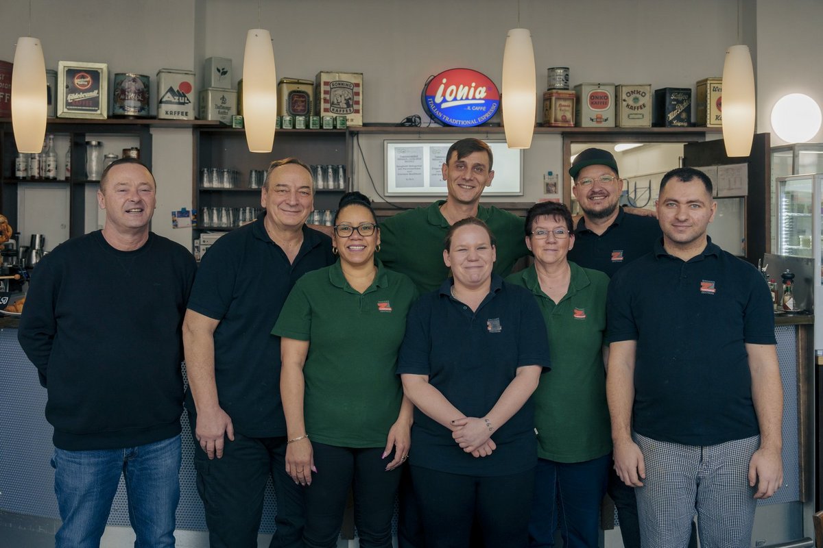 Lächelnde Mitarbeiter in grünen und dunklen Poloshirts posieren vor einer Bartheke. Im Hintergrund sind Regale mit Dekorationsgegenständen und ein Leuchtschild zu sehen.