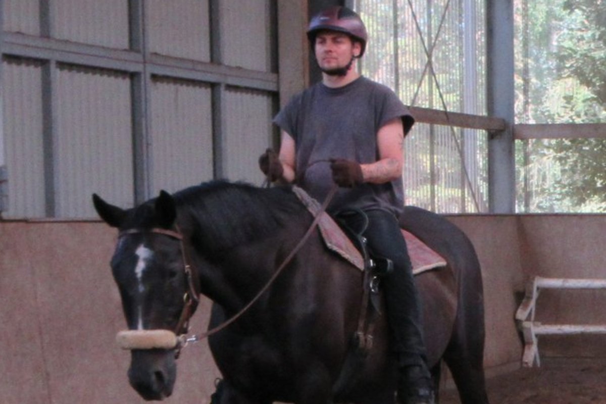 Ein Mann mit Helm und grauem T-Shirt reitet ein schwarzes Pferd mit weißem Stirnbrand in einer Reithalle. Das Pferd hat ein weißes Hinterbein und bewegt sich auf dem Sandboden.