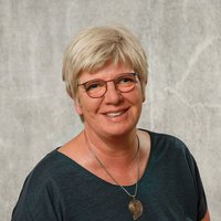 Porträtaufnahme einer lächelnden Frau mit kurzen blonden Haaren und Brille. Sie trägt ein dunkelgrünes T-Shirt und eine Halskette vor einer strukturierten grauen Wand.