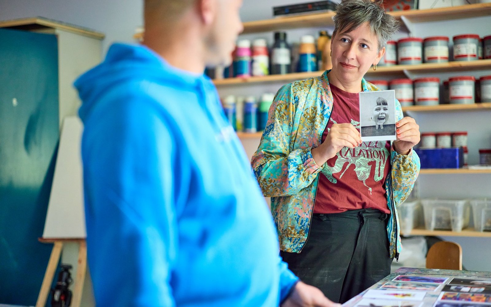 Eine Frau mit kurzen Haaren und einer bunten Jacke hält ein Foto hoch und zeigt es einem Mann im blauen Hoodie. Im Hintergrund sind Regale mit Farbdosen zu sehen.