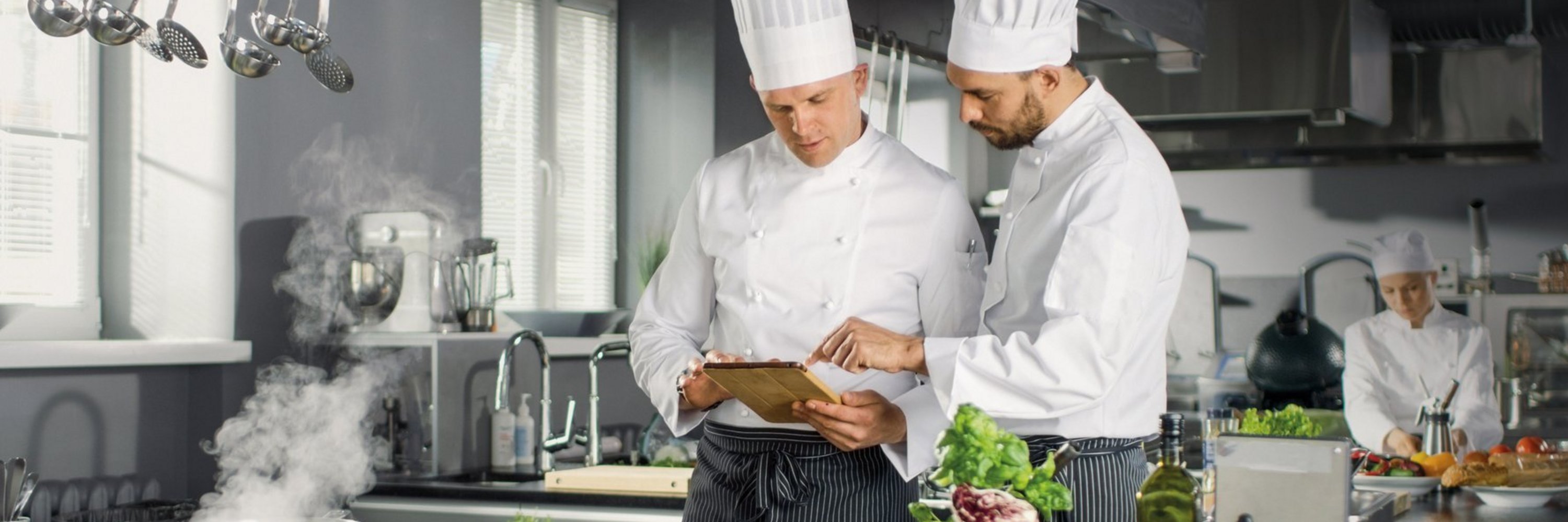 Zwei Köche in weißen Uniformen stehen in einer professionellen Küche und diskutieren über einen Plan auf einem Tablet, wobei einer auf den Bildschirm zeigt. Im Vordergrund liegen frische Zutaten und dampfende Töpfe.