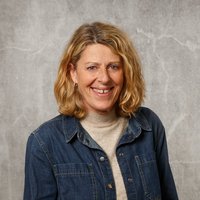 Eine lächelnde Frau mit schulterlangen blonden Haaren steht vor einer grauen, texturierten Wand. Sie trägt eine blaue Jeansjacke über einem beigen Pullover.