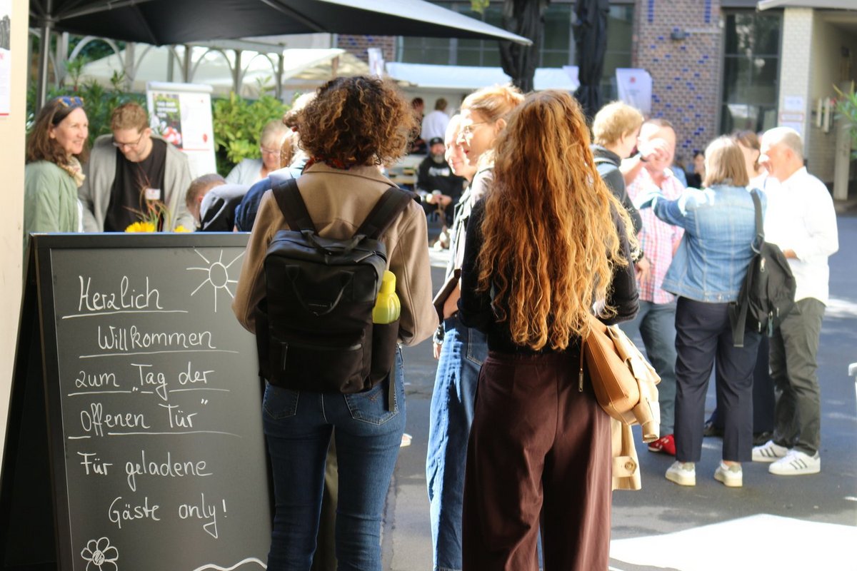 Ein Schild im Vordergrund begrüßt mit der Aufschrift „Herzlich Willkommen zum Tag der offenen Tür“ für geladene Gäste. Dahinter unterhalten sich Gäste auf einer Außenveranstaltung, wobei zwei Frauen im Fokus stehen.