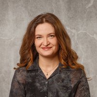 Eine lächelnde junge Frau mit langen braunen Haaren steht mit verschränkten Armen vor einer grauen Wand und trägt eine dunkle, durchscheinende Bluse.