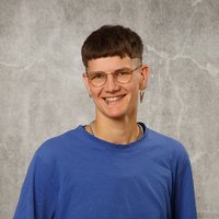 Ein junger Mann mit kurzen Haaren und Brille lächelt freundlich in die Kamera. Er trägt ein blaues T-Shirt und steht vor einem grauen, strukturierten Hintergrund.