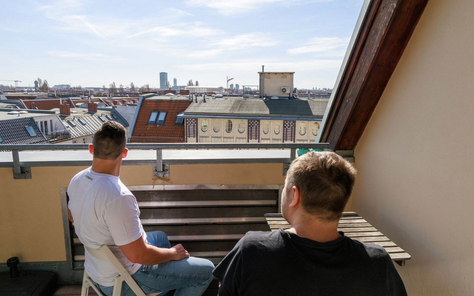 Zwei Männer sitzen auf einem Balkon und blicken auf eine städtische Skyline. Links sitzt einer im weißen T-Shirt auf einem Stuhl, rechts einer im schwarzen auf einer Bank. Im Hintergrund sind Dächer und ein Turm unter blauem Himmel zu sehen.