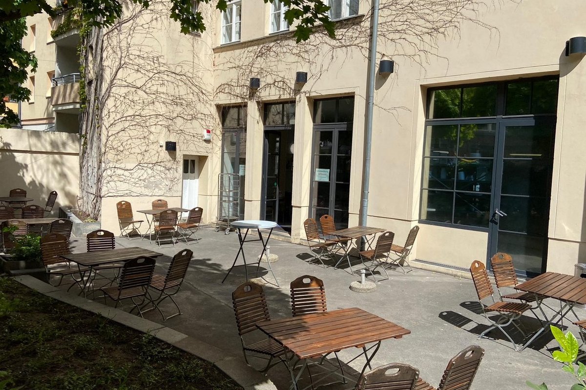 Eine sonnige Außenterrasse mit Holztischen und Stühlen auf einem gepflasterten Hof. Im Hintergrund steht ein hellgelbes Gebäude mit großen Glasfenstern und Türen. An der Fassade ranken sich Pflanzen.