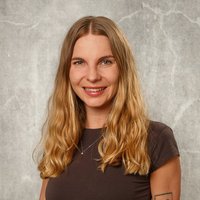 Porträt einer lächelnden jungen Frau mit langen blonden Haaren und Tattoos an den Armen, die lässig vor einer grauen Betonwand steht. Sie trägt ein dunkles T-Shirt.