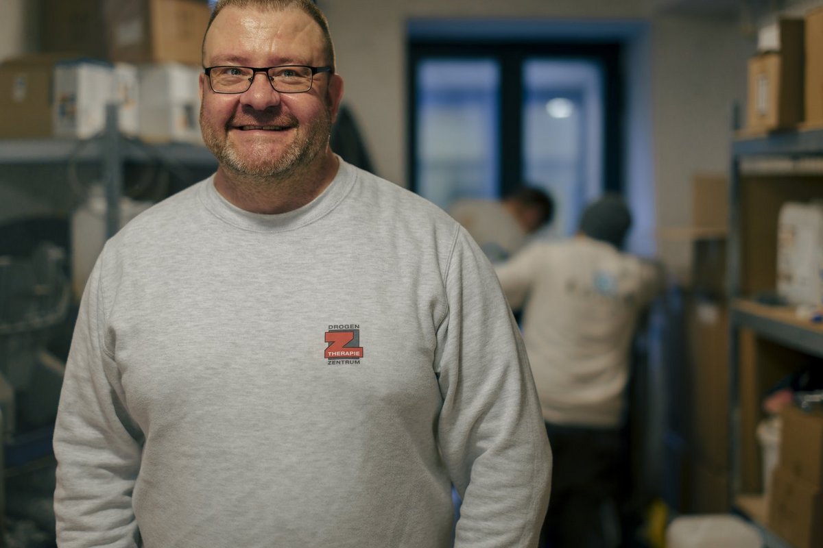 Ein lächelnder Mann mit Brille und grauem Sweatshirt steht in einer Lagerhalle. Im unscharfen Hintergrund sind Regale mit Kartons sowie weitere Mitarbeiter zu erkennen.