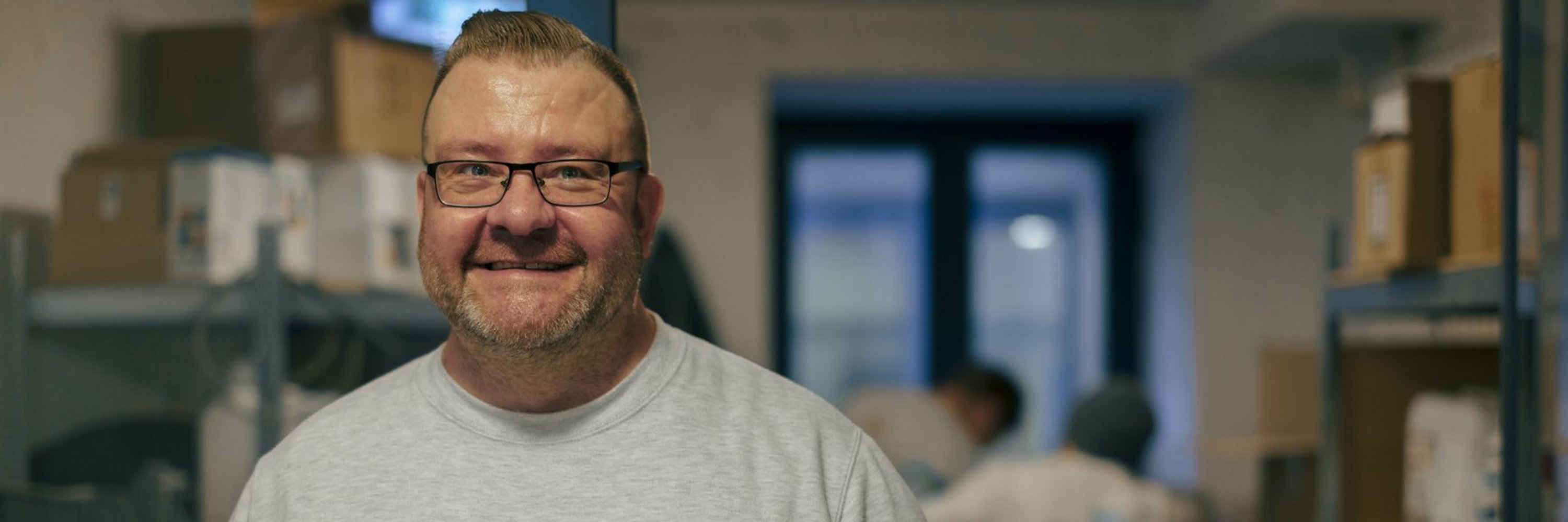 Ein lächelnder Mann mit Brille und grauem Sweatshirt steht in einer Lagerhalle. Im unscharfen Hintergrund sind Regale mit Kartons sowie weitere Mitarbeiter zu erkennen.