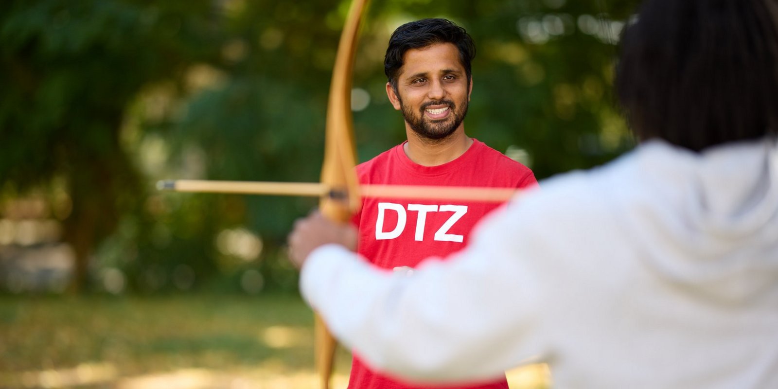 Ein lächelnder Mann in einem roten T-Shirt mit der Aufschrift „DTZ“ hält einen Bogen und Pfeil und zielt auf eine Person im weißen Hoodie im Vordergrund. Der Hintergrund ist ein grüner, unscharfer Park.