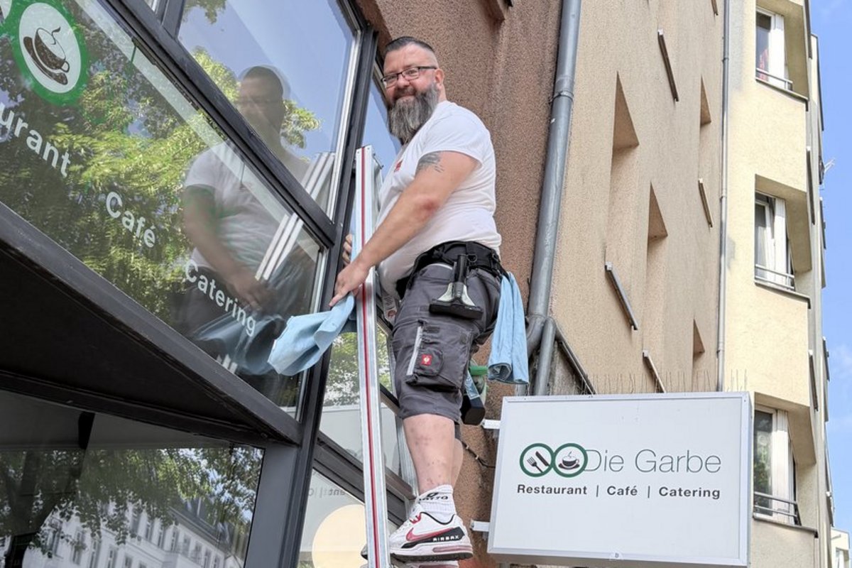 Ein Mann mit Bart und Brille steht auf einer Leiter und putzt das Fenster des Restaurants „Die Garbe". Er hält ein blaues Tuch in der Hand.
