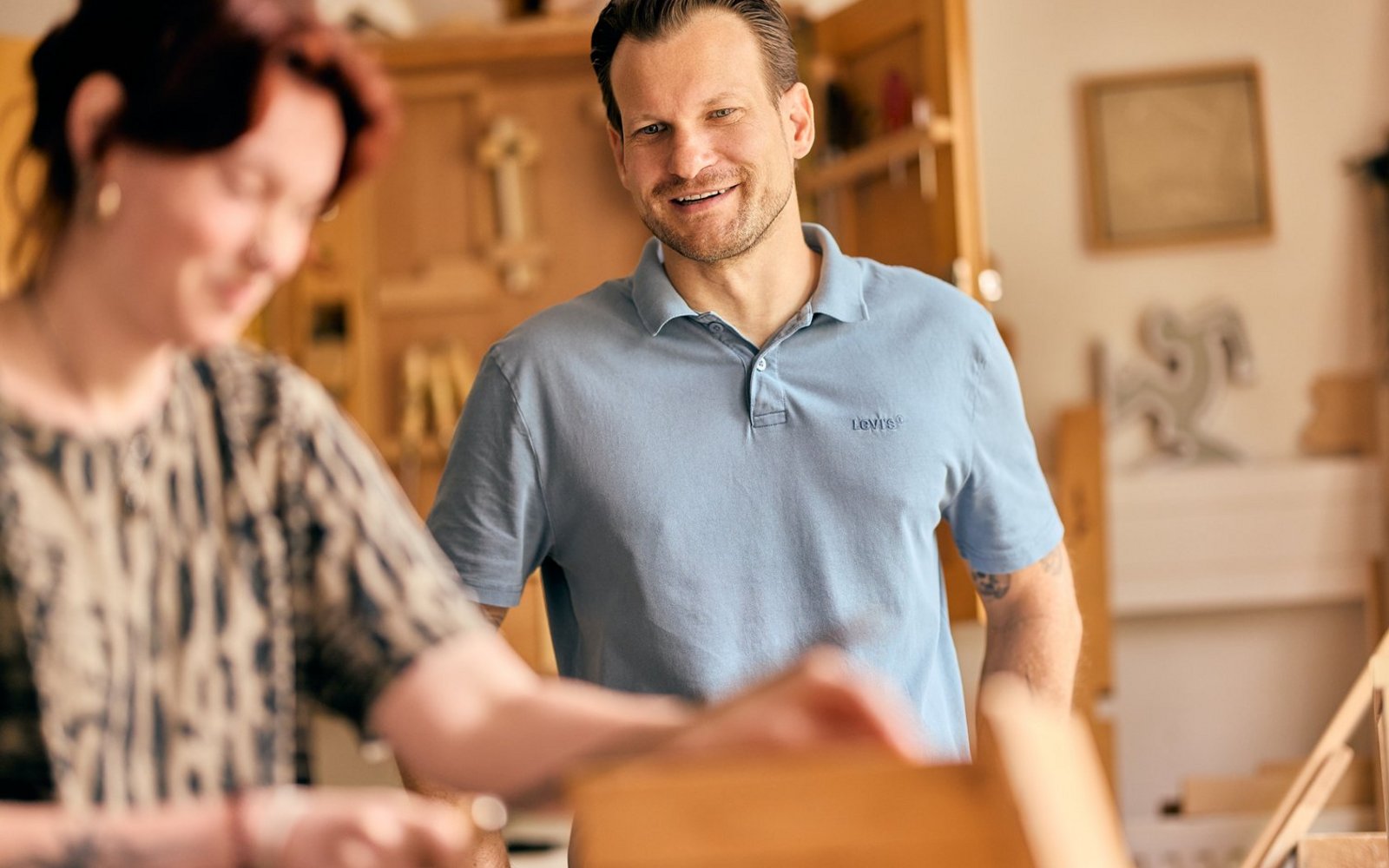 Ein Mann in einem hellblauen Poloshirt steht in einer Holzwerkstatt und lächelt freundlich in die Kamera. Im unscharfen Vordergrund arbeitet eine Frau an einem Holzstück. Im Hintergrund sind Werkbankmöbel und Werkzeuge zu erkennen.