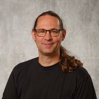 Ein lächelnder Mann mit Brille und langem, zurückgebundenem Haar steht vor einem grauen Hintergrund. Er trägt ein schwarzes T-Shirt.