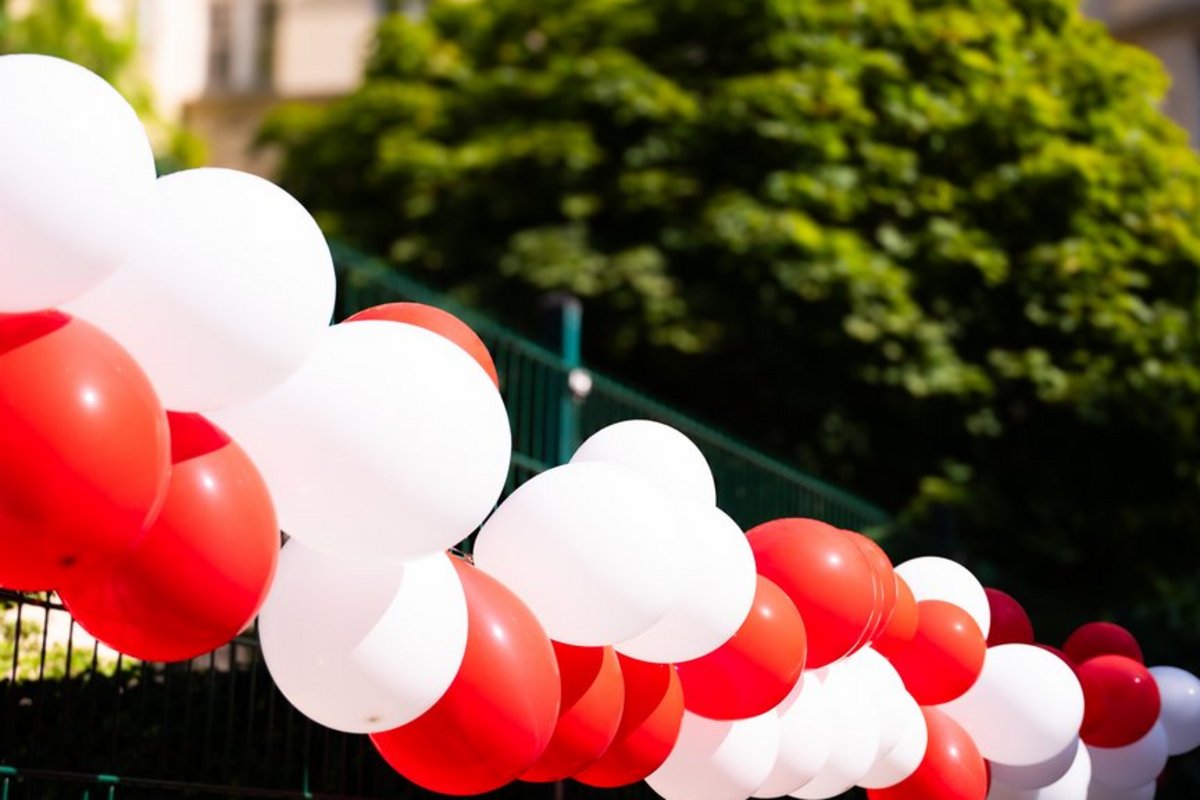 Abwechselnd rote und weiße Luftballons sind an einem grünen Zaun befestigt. Im unscharfen Hintergrund sind Bäume und ein Gebäude zu erkennen.